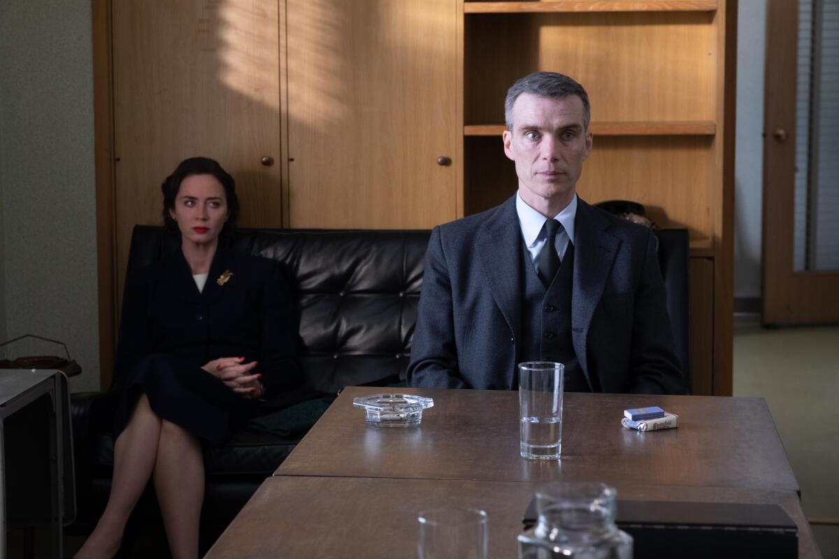 Cillian Murphy sits straight and tall at a conference table while Emily Blunt sits behind him on a couch in "Oppenheimer."