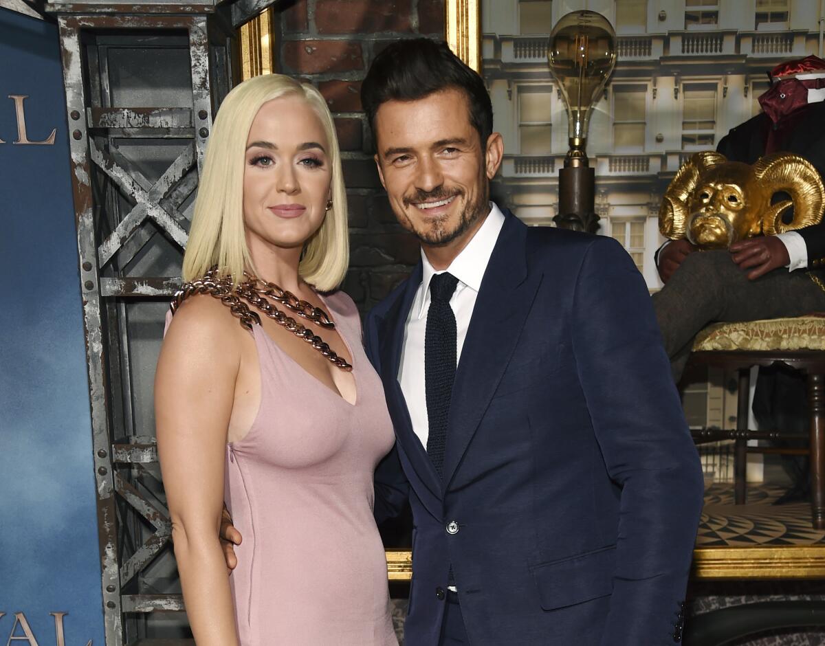 Katy Perry wears a mauve dress while standing with Orlando Bloom, clad in a navy suit 