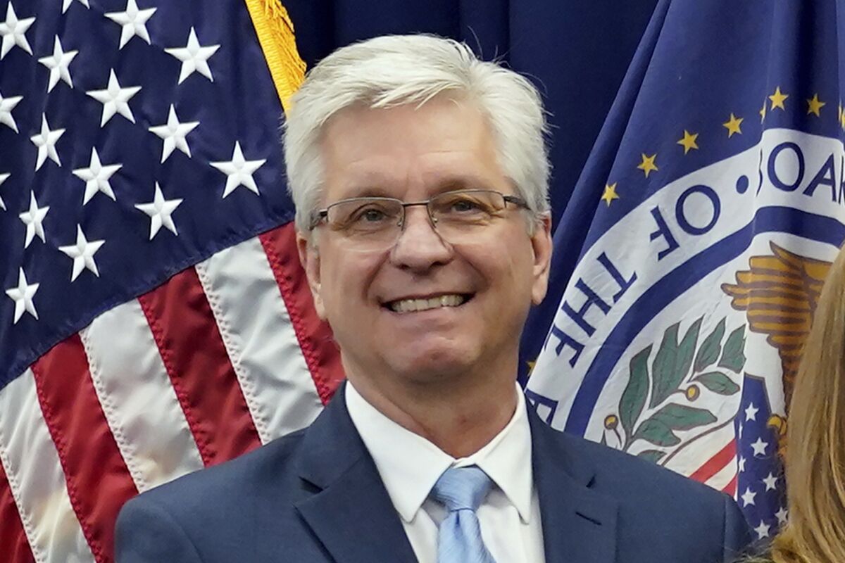 A man wearing glasses smiles while posing in front of two flags.
