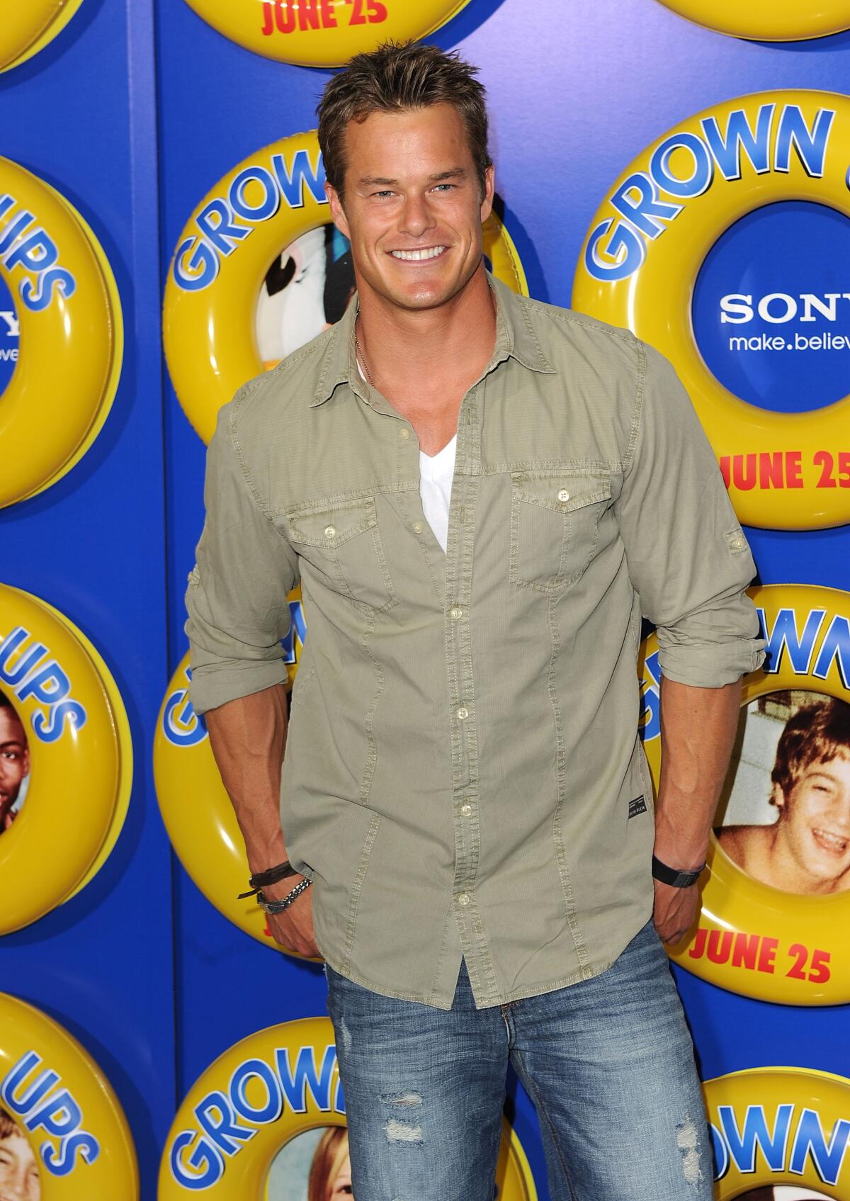Alec Musser wears a khaki button-down shirt over a white shirt and blue jeans as he poses for photos in front of a backdrop