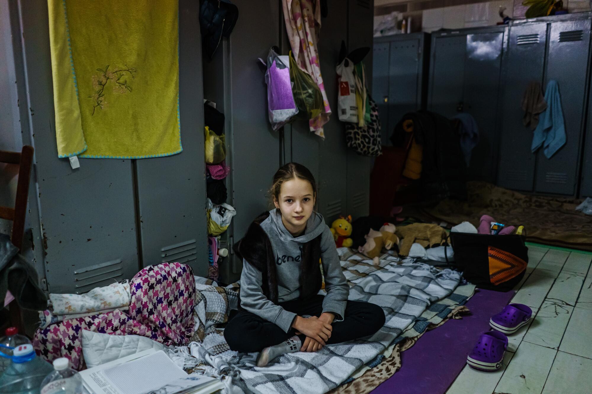 A 10-year-old girl wearing a gray long-sleeved shirt sits on top of blankets on a tiled floor next to metal storage lockers.