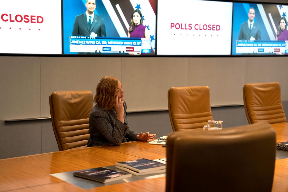 Sarah Snook as Shiv Roy sits at a conference table beneath television screens while talking on the phone.