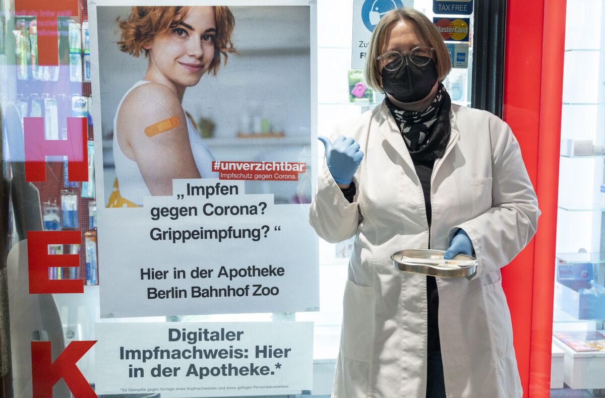 Sandra Fundel, pharmacist at the pharmacy at Bahnhof Zoo, stands outside the pharmacy with vaccination paraphernalia, next to a poster advertising the Corona vaccination in Berlin, Germany, Monday, Feb. 7, 2022. Pharmacists in the country are scheduled to begin offering vaccinations Tuesday, after parliament changed the rules so that they, dentists and vets could administer the shoots. (Christophe Gateau/dpa via AP)