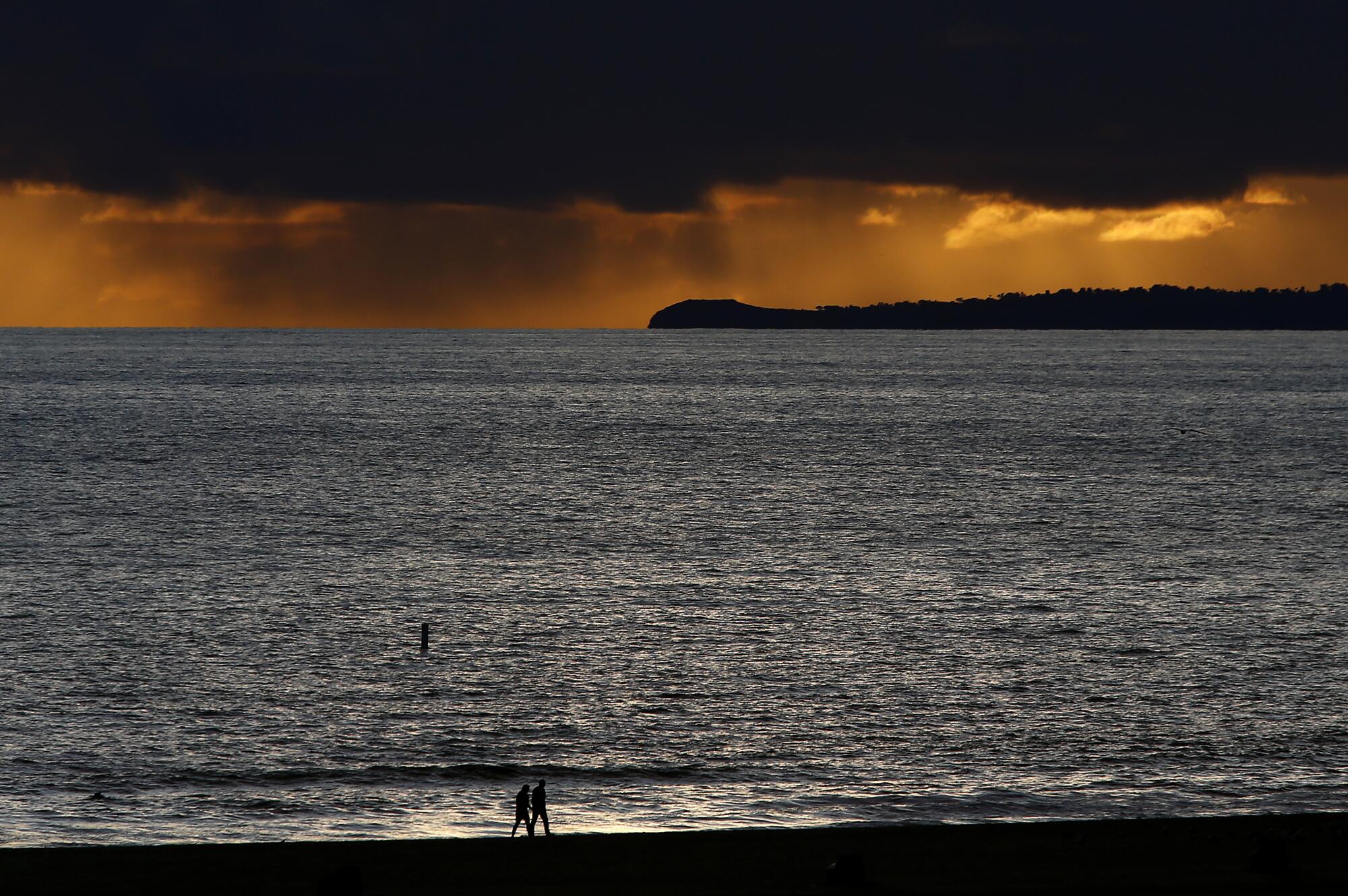 A couple walks alone on Santa Monica Beach as rain falls from storm clouds in the distance.