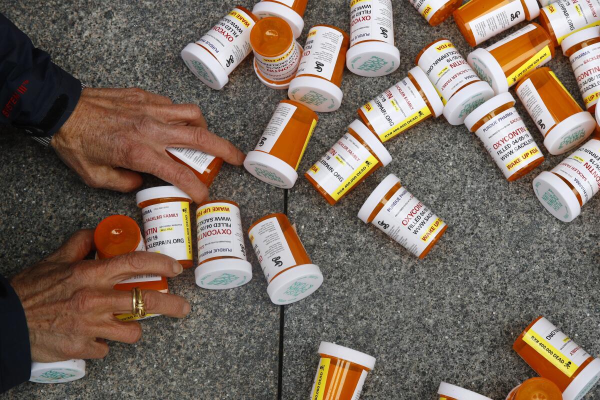 A pair of hands grabbing some of over two dozen pill bottles lying on a sidewalk, with the word "OxyContin" visible on some.