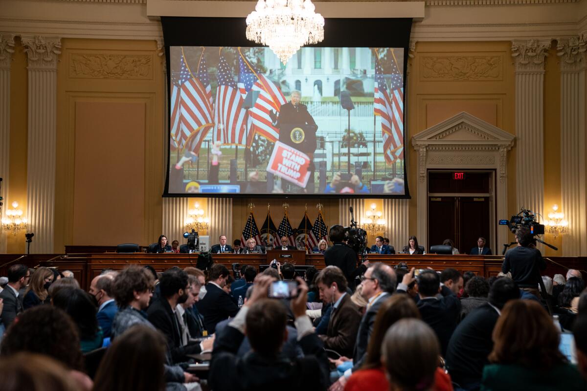 An image of then-President Trump at his Jan. 6 rally is displayed on a large screen above lawmakers leading a crowded hearing