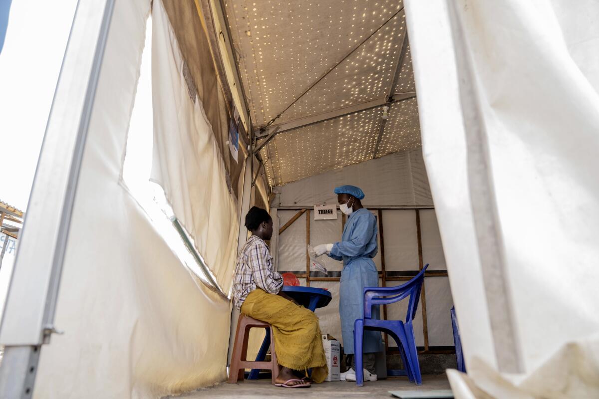 A patient sits for care in a tent next to a standing worker wearing scrubs and a mask.