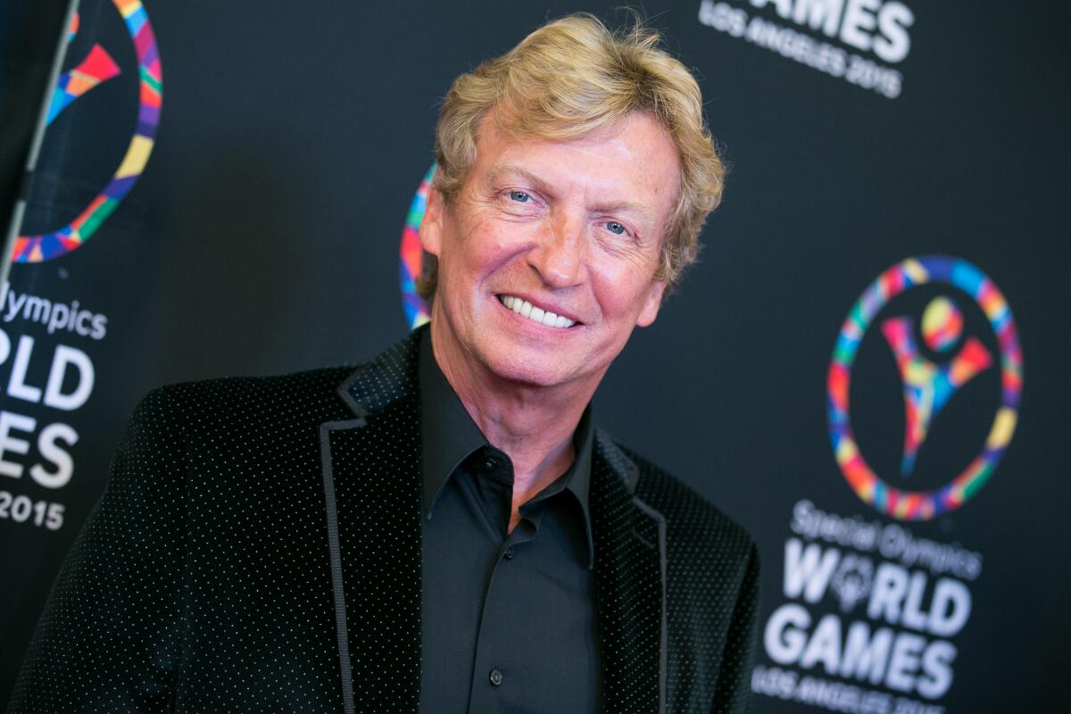 Nigel Lythgoe, with dirty blond hair, smiles while posing in a black suit jacket and black dress shirt 
