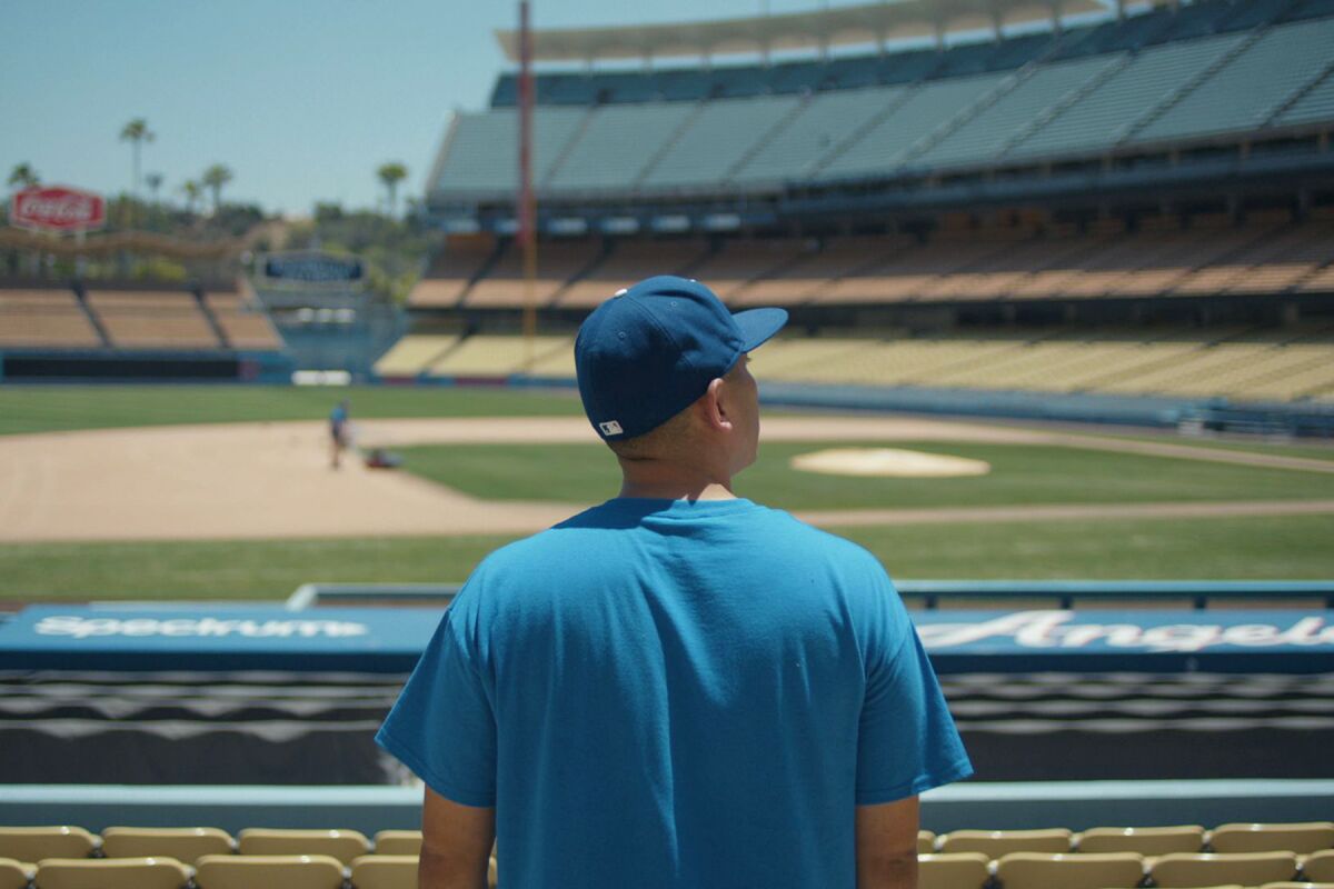 A man in a blue T-shirt and baseball cap looks out from the stands at the infield in an empty Dodger Stadium.