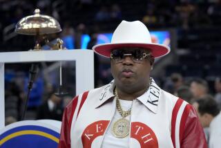 E-40 wears sunglasses, a white-and-red jacket and a white hat inside a basketball arena