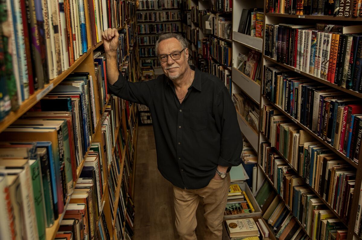 A man stands between rows of bookshelves, his upraised right arm resting against a shelf.