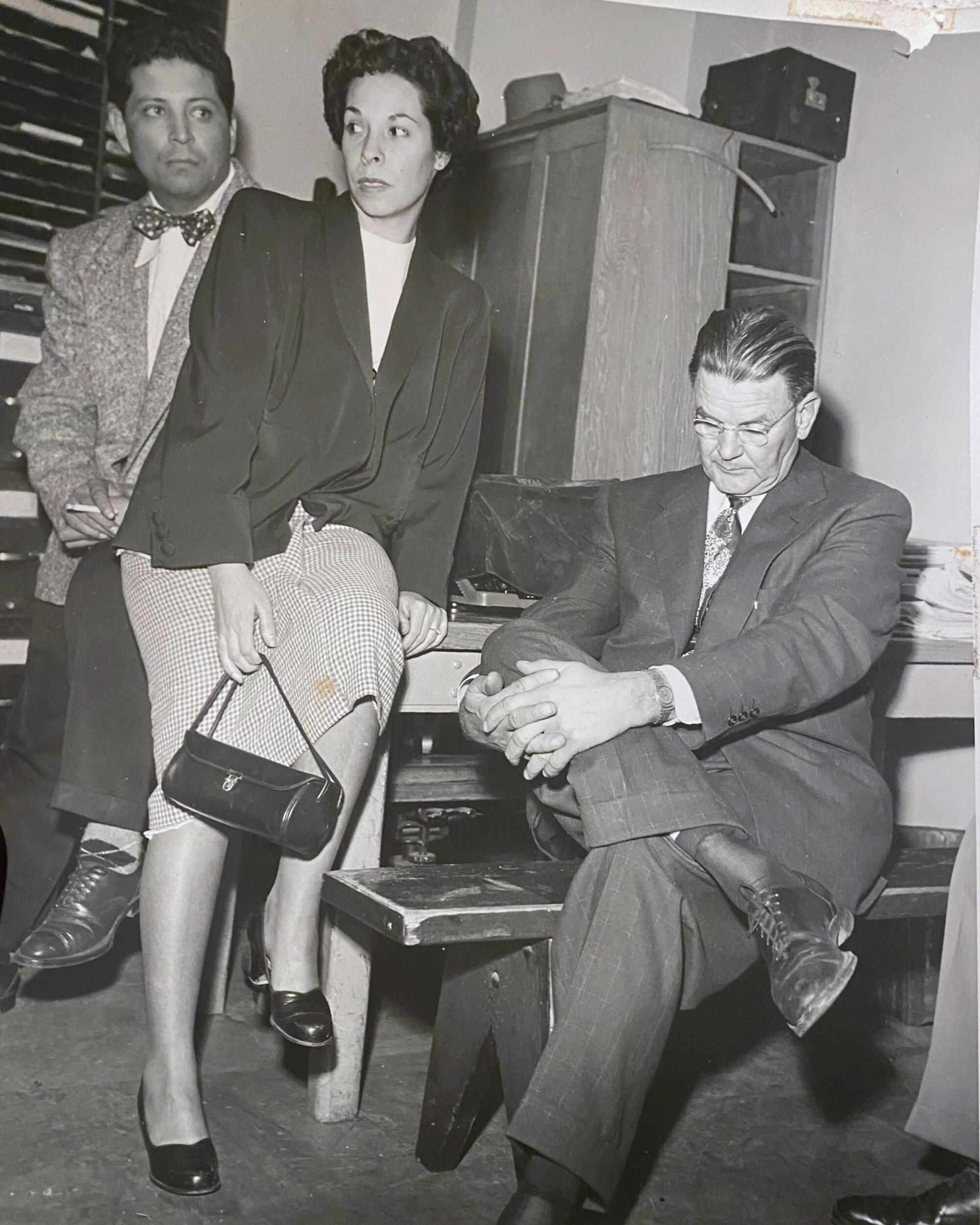 Three people at a table. A man in bowtie and suit, a woman with dark hair, blazer and skirt, a man in suit looking downward.