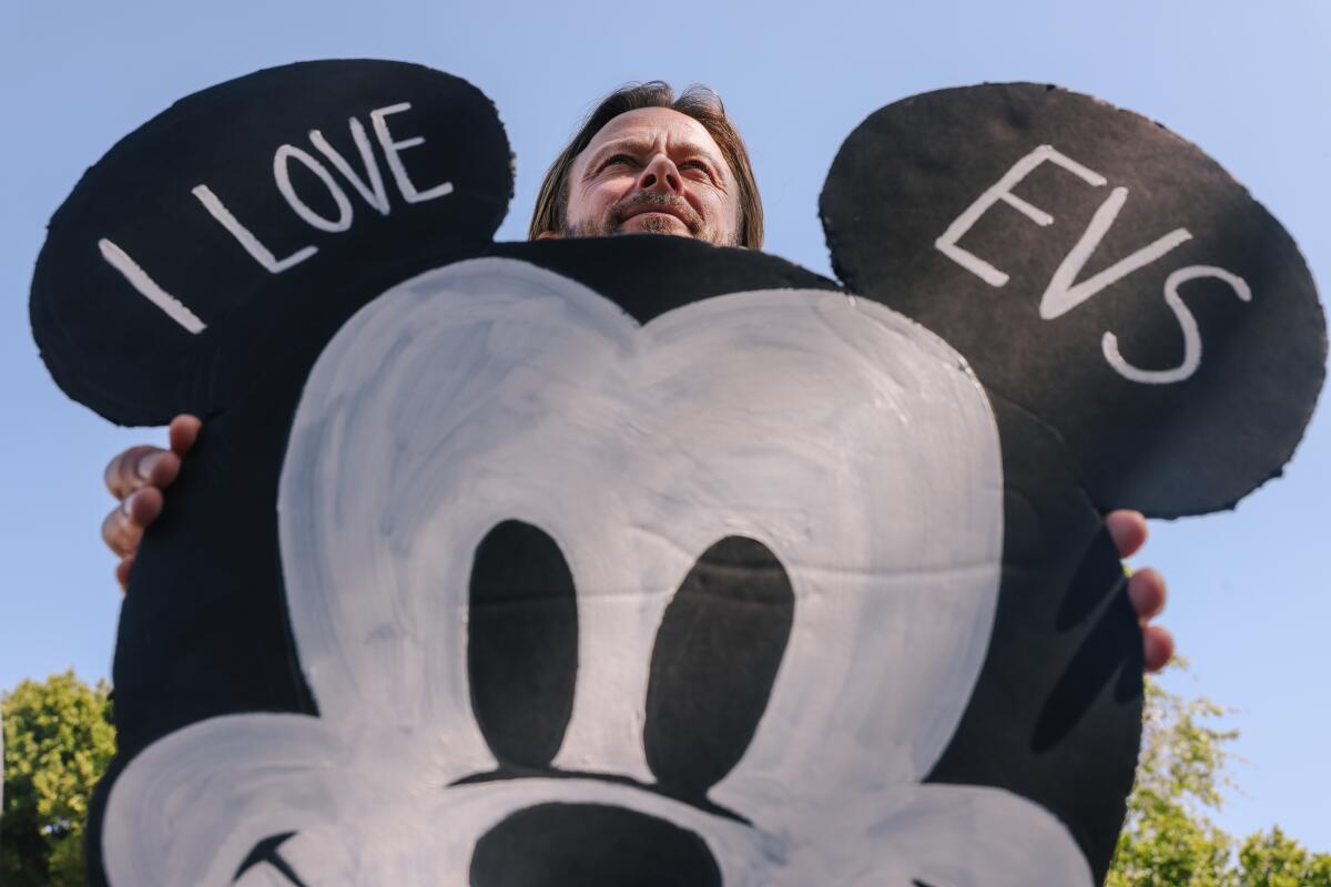 A man holds a Mickey Mouse-shaped sign that reads "I love EVs." 