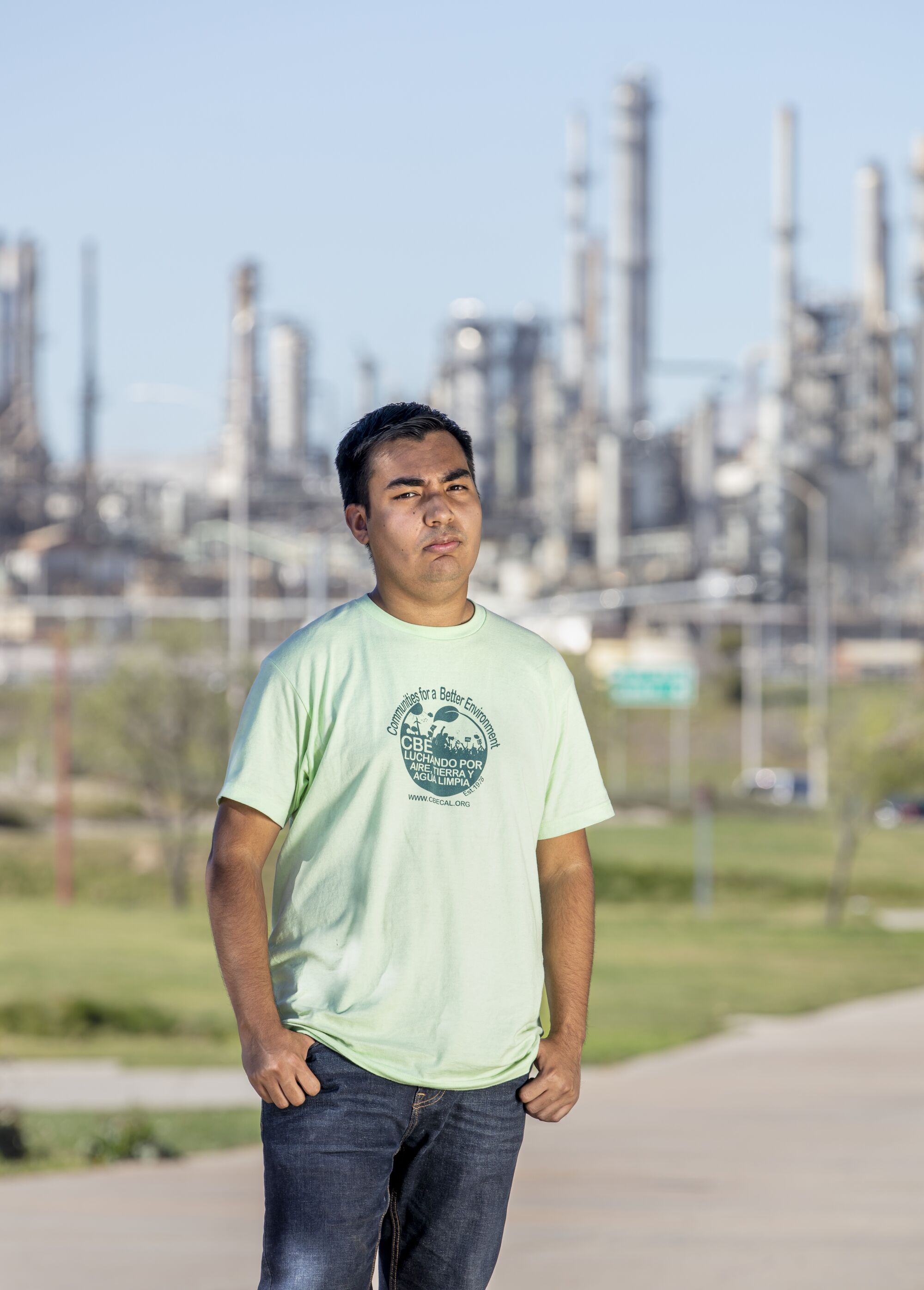 A young man stands with an oil refinery in the background