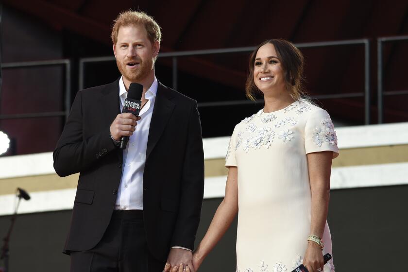 Prince Harry in a black blazer and pants standing next to and holding hands with Meghan Markle in a white dress 