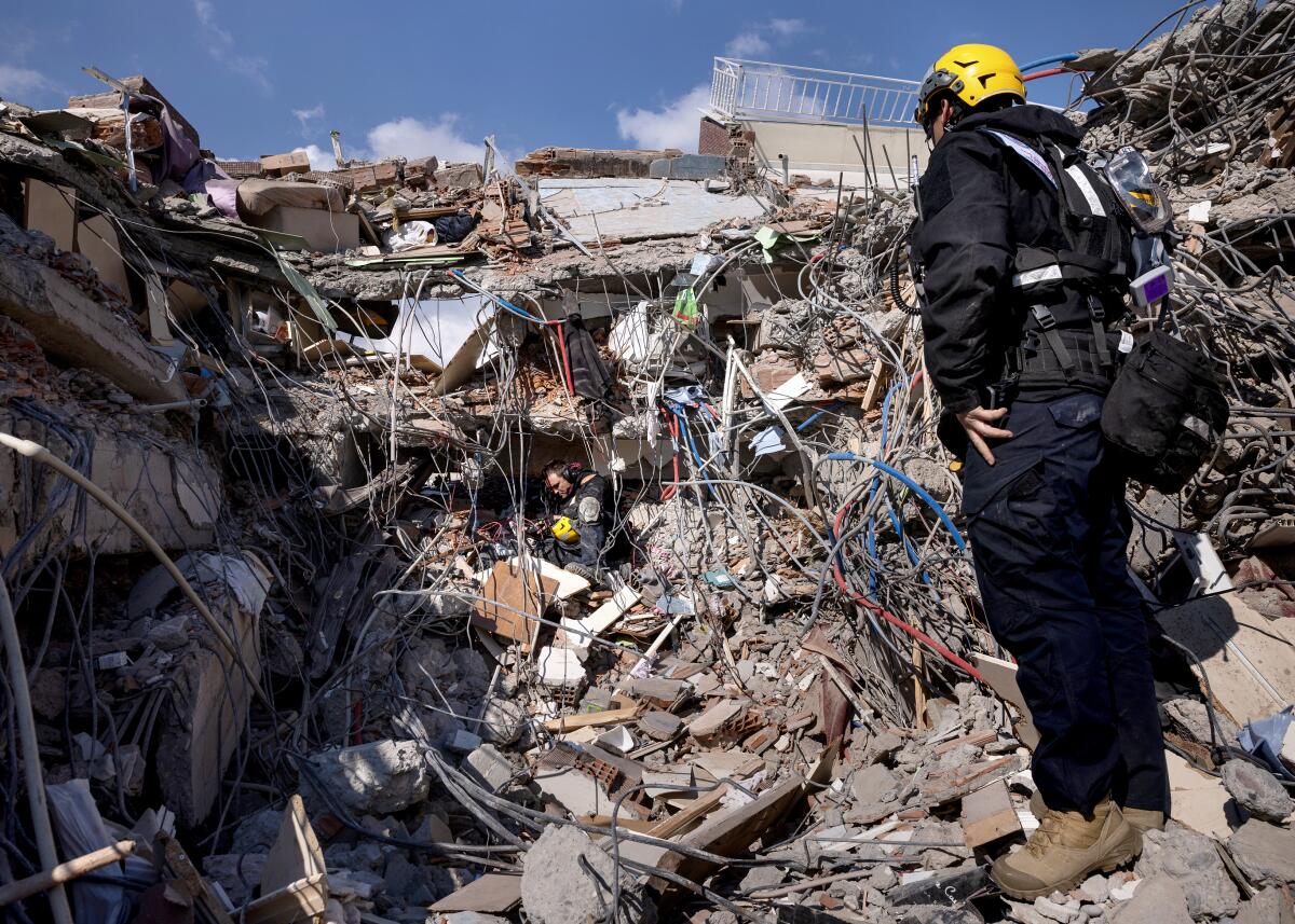A man wearing headphones and holding a yellow helmet sits in a nest of rubble.