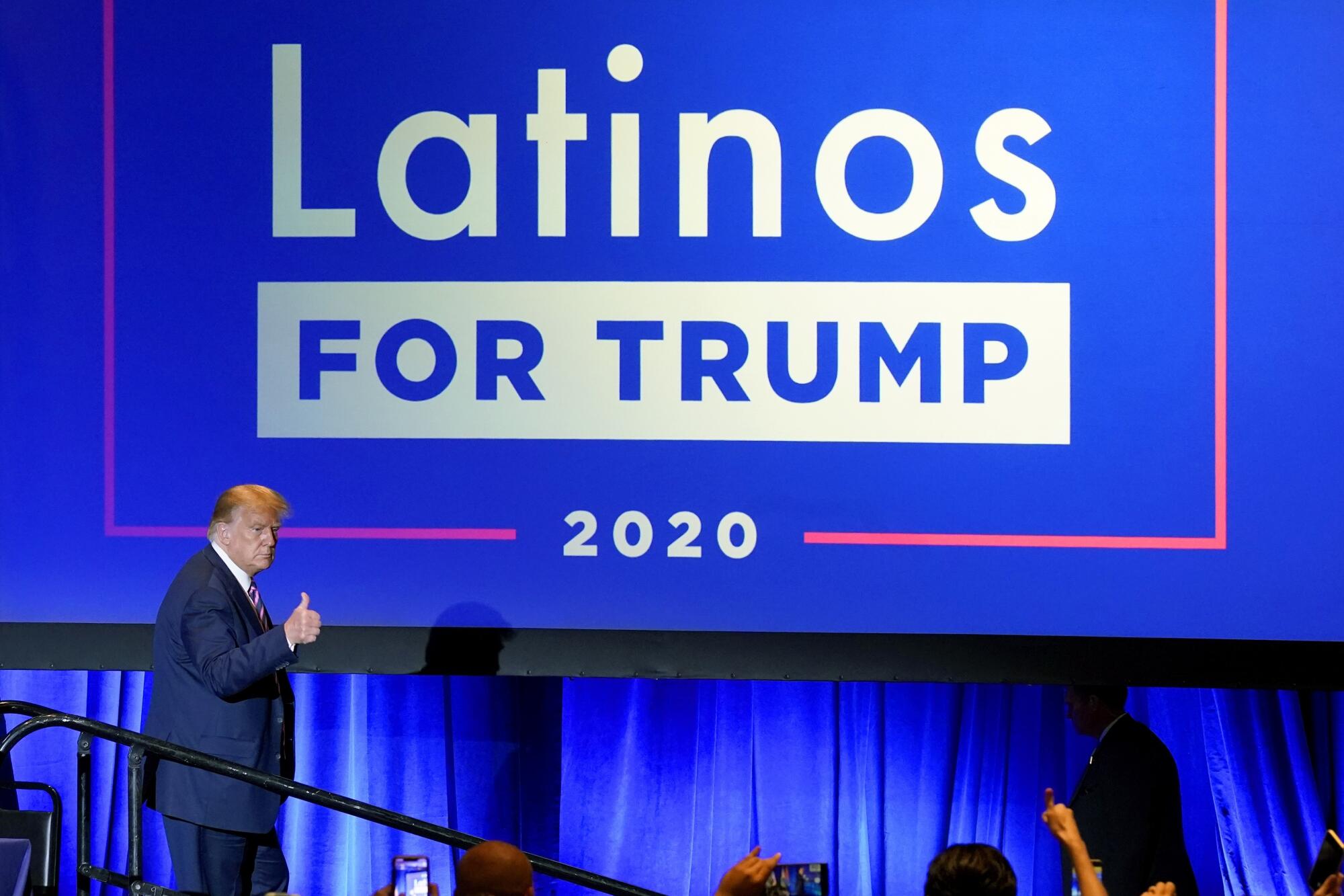President Trump pictured from a distance below a large blue "Latinos for Trump 2020" sign, giving a thumbs-up toward a crowd