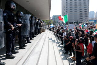 Law enforcement officers atop steps at the front of a building face a crowd at the bottom of the steps.