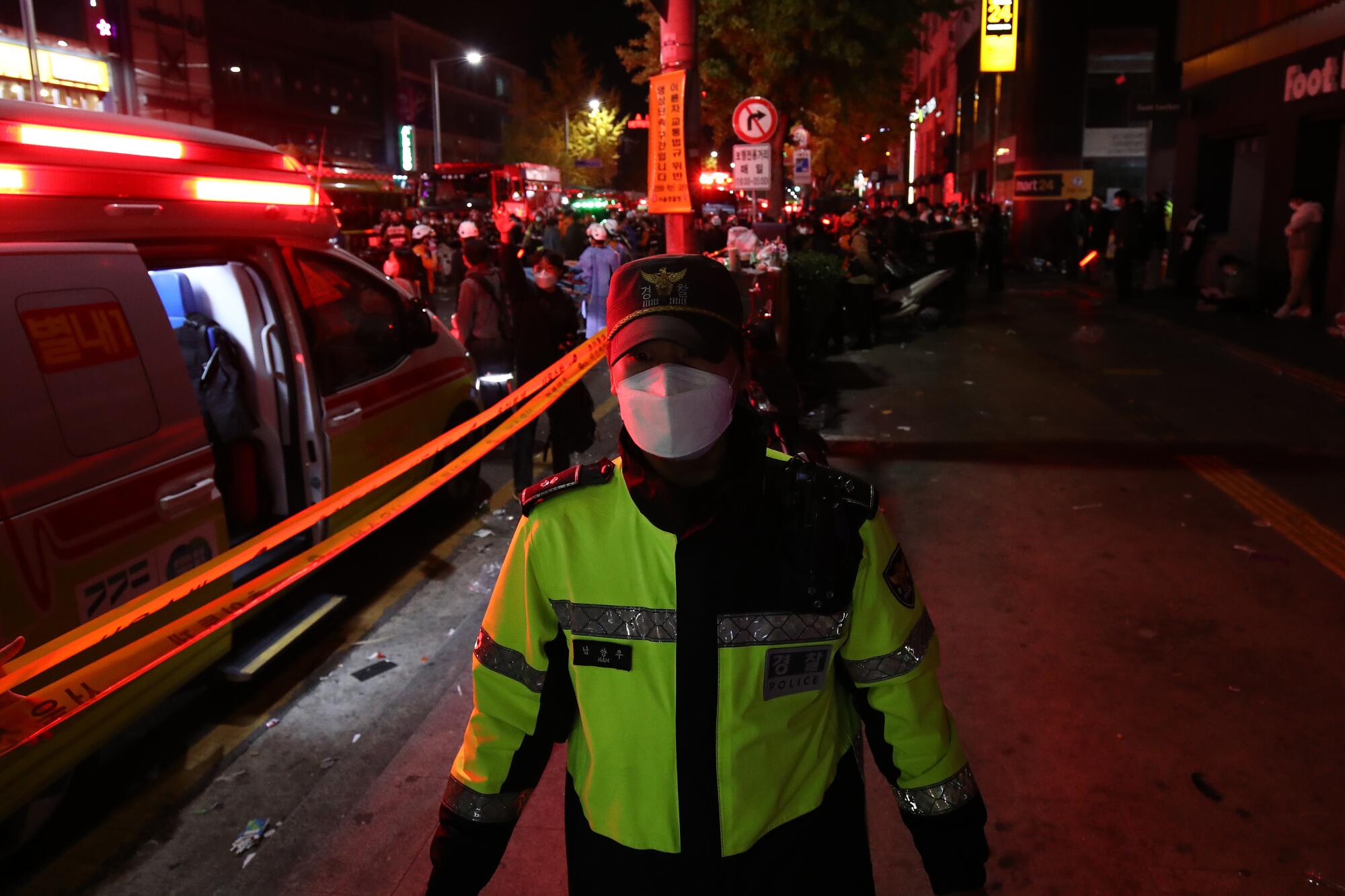 A police officer in a yellow-green vest is surrounded by the red glow of lights on a crowded street with emergency vehicles