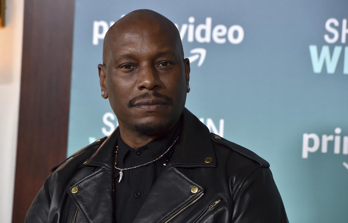 Tyrese Gibson poses in a black shirt, black leather jacket and chain against a blue backdrop.