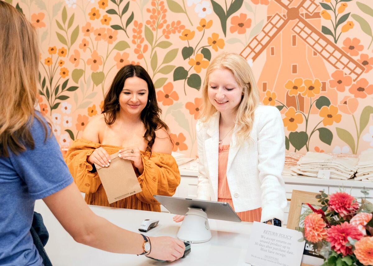 Two women stand behind a store counter while another woman prepares to pay.