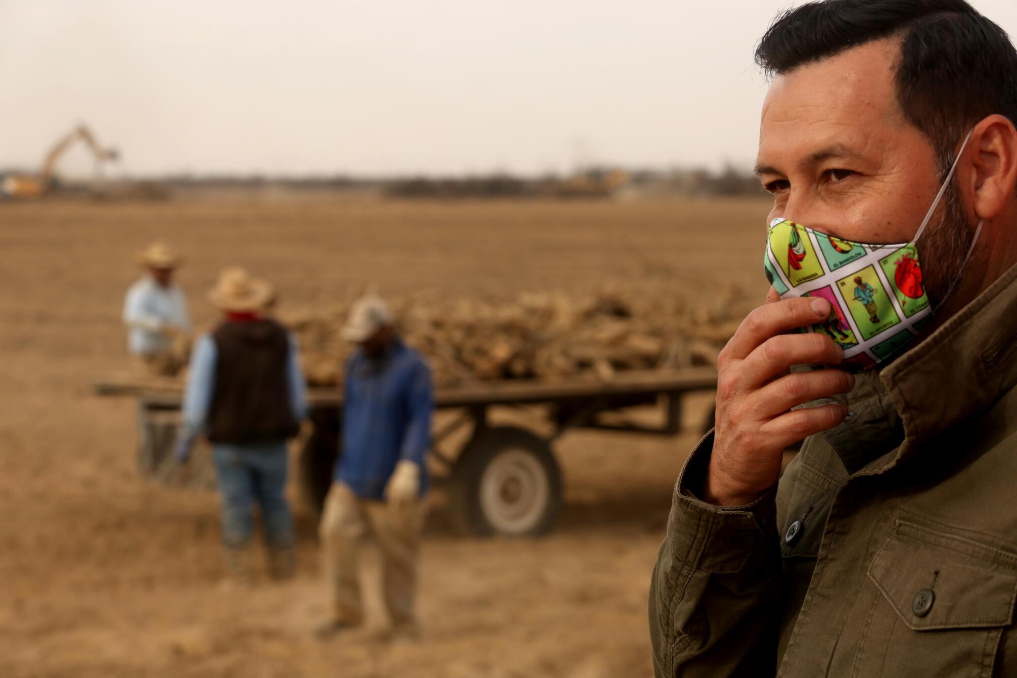 Ricardo Castorena stands with his hand touching his mask as farmworkers work in the background