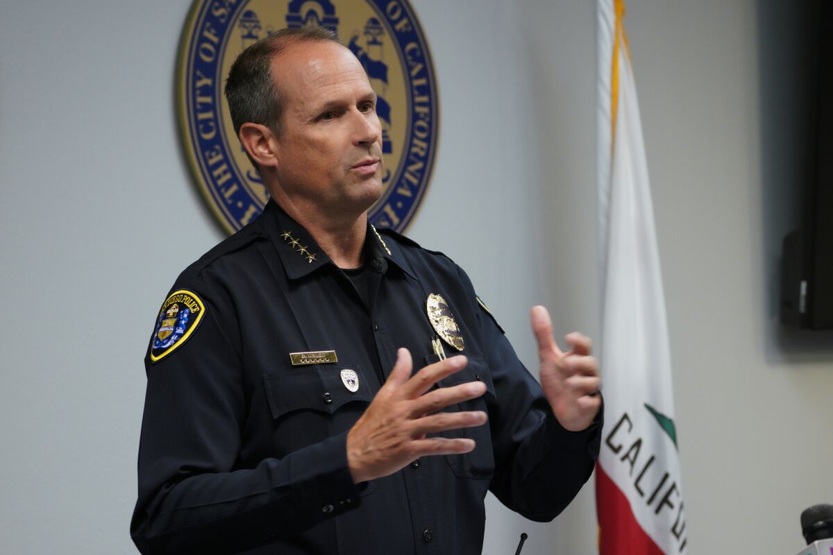 A man wearing a police uniform gestures as he speaks at a press conference next to a California state flag.
