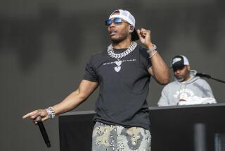 Nelly in a black t-shirt, chains, a backwards cap and camoflauge-patterned pants performing on a stage