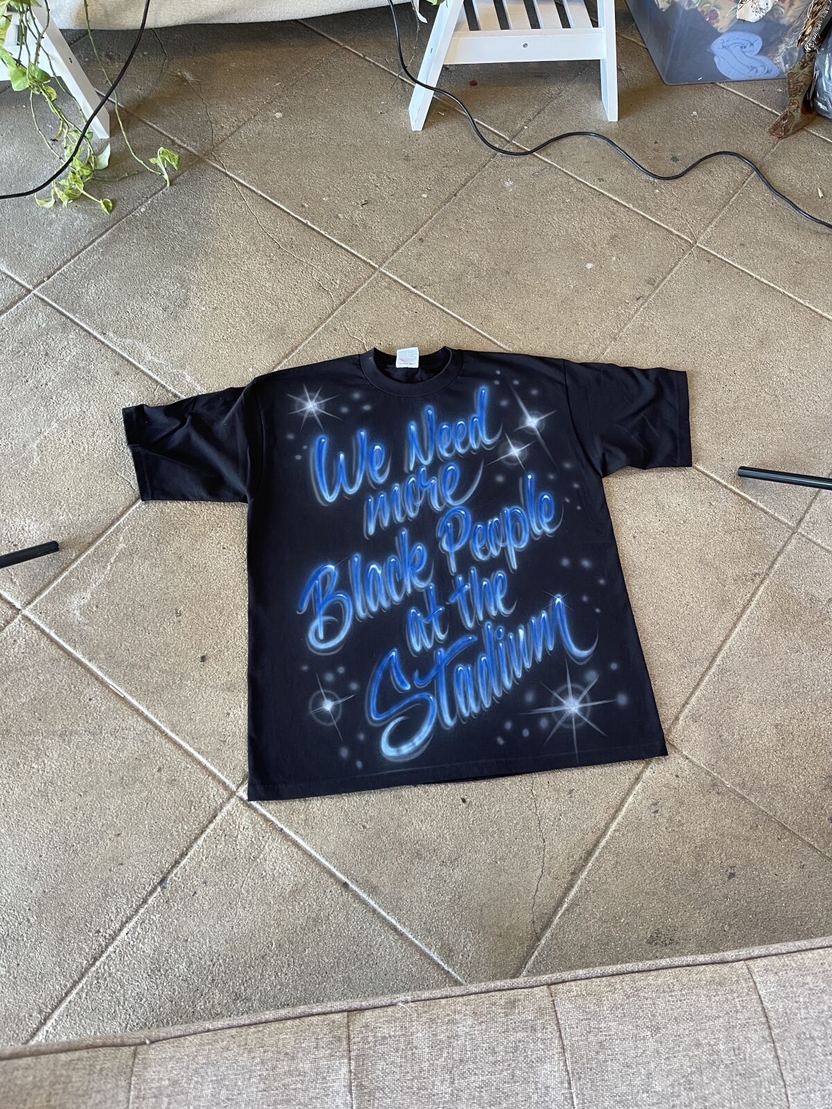A black T-shirt with the words "We Need more Black People at the Stadium" airbrushed in blue and white