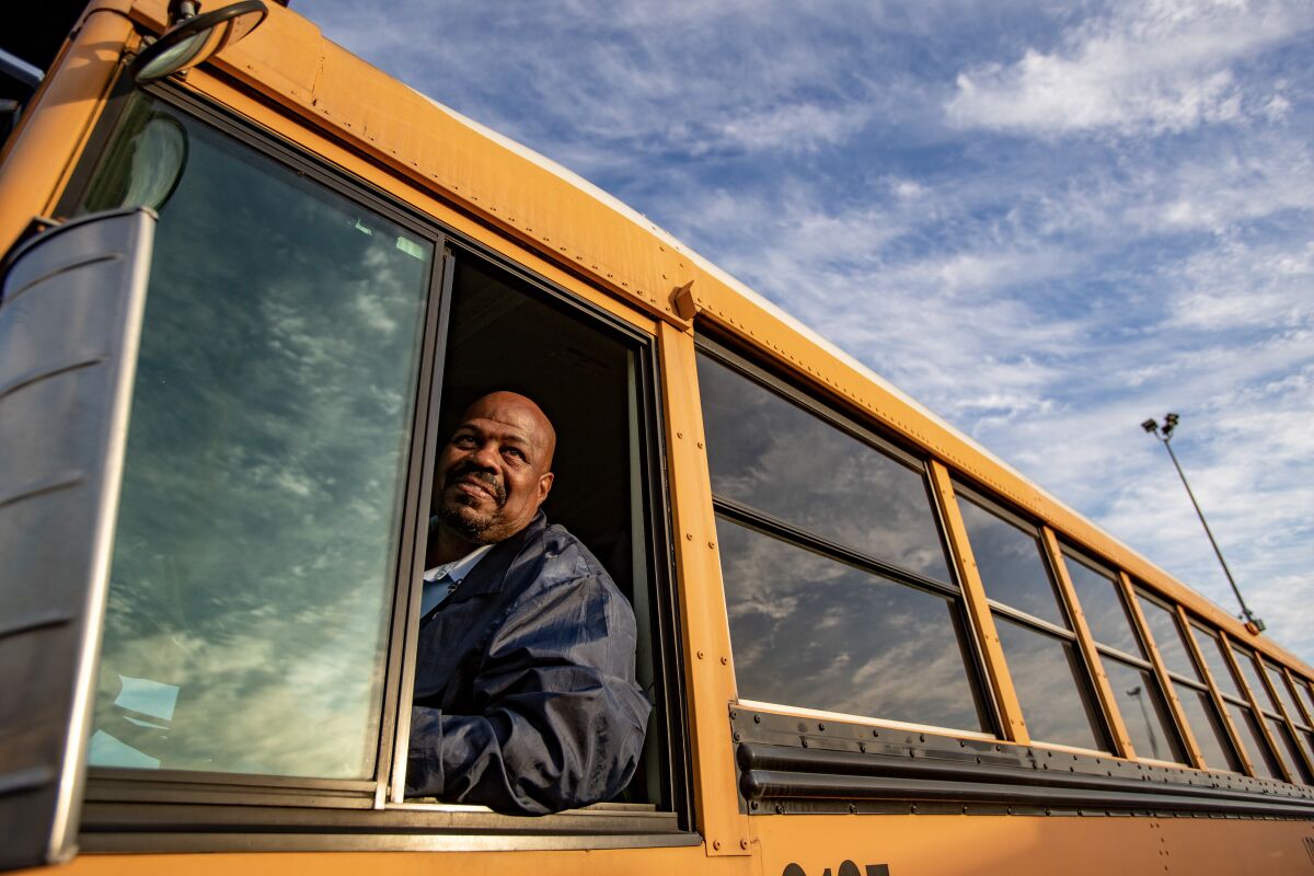 A older Black man with a shaved head and blue jacket sits in the drivers seat of a school bus