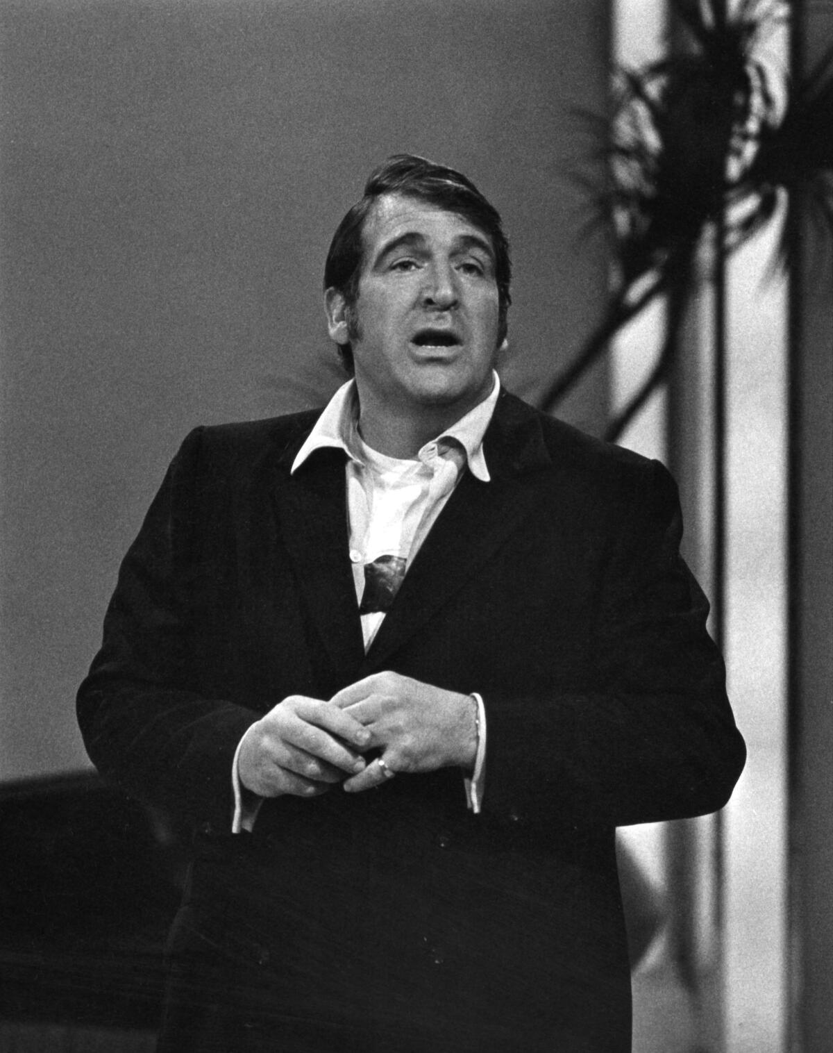 Shecky Greene wears a black blazer with a white collared shirt as he talks on a stage.
