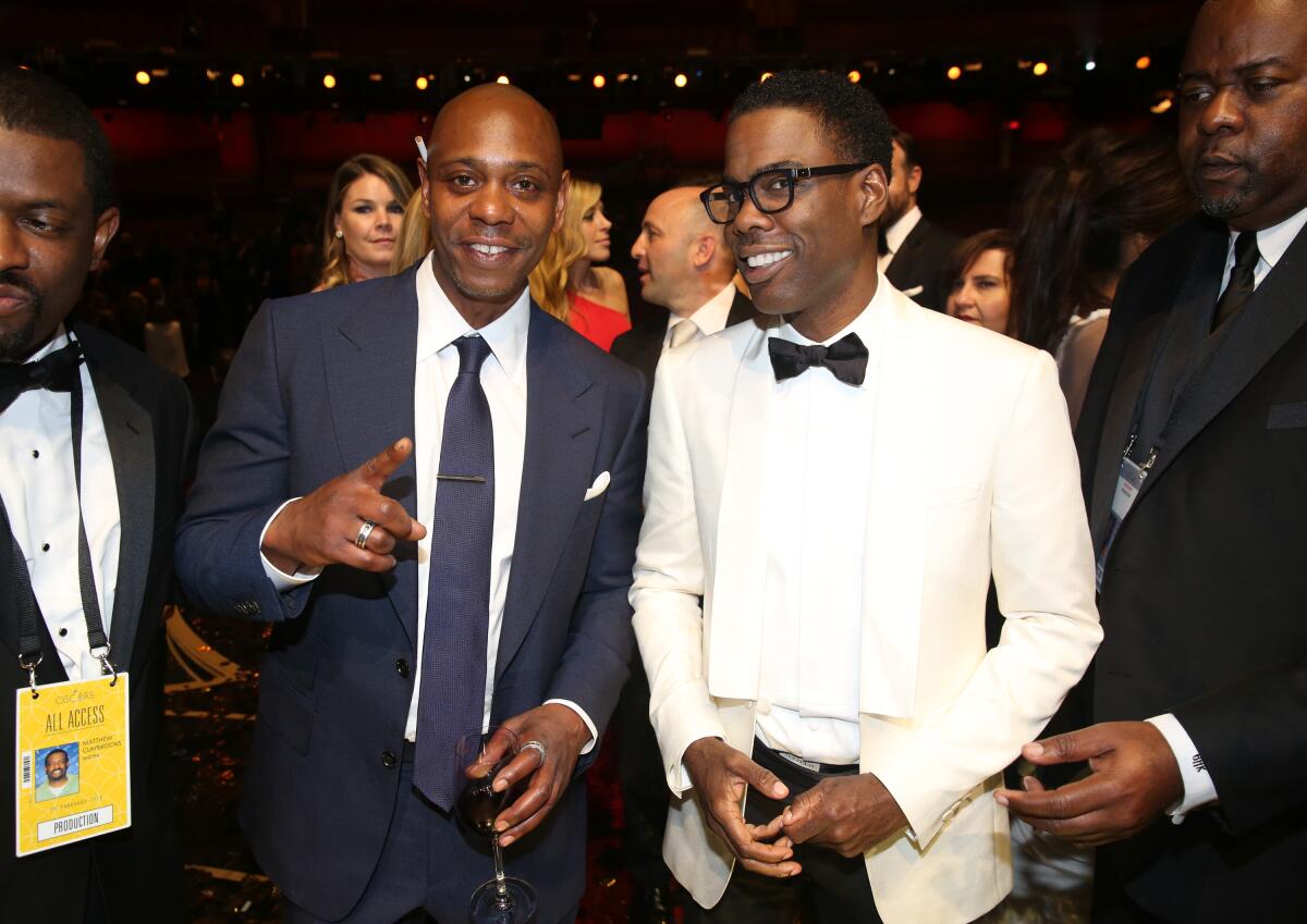 A man in a blue suit stands next to a man in a white suit and black bow tie, and the two smile.