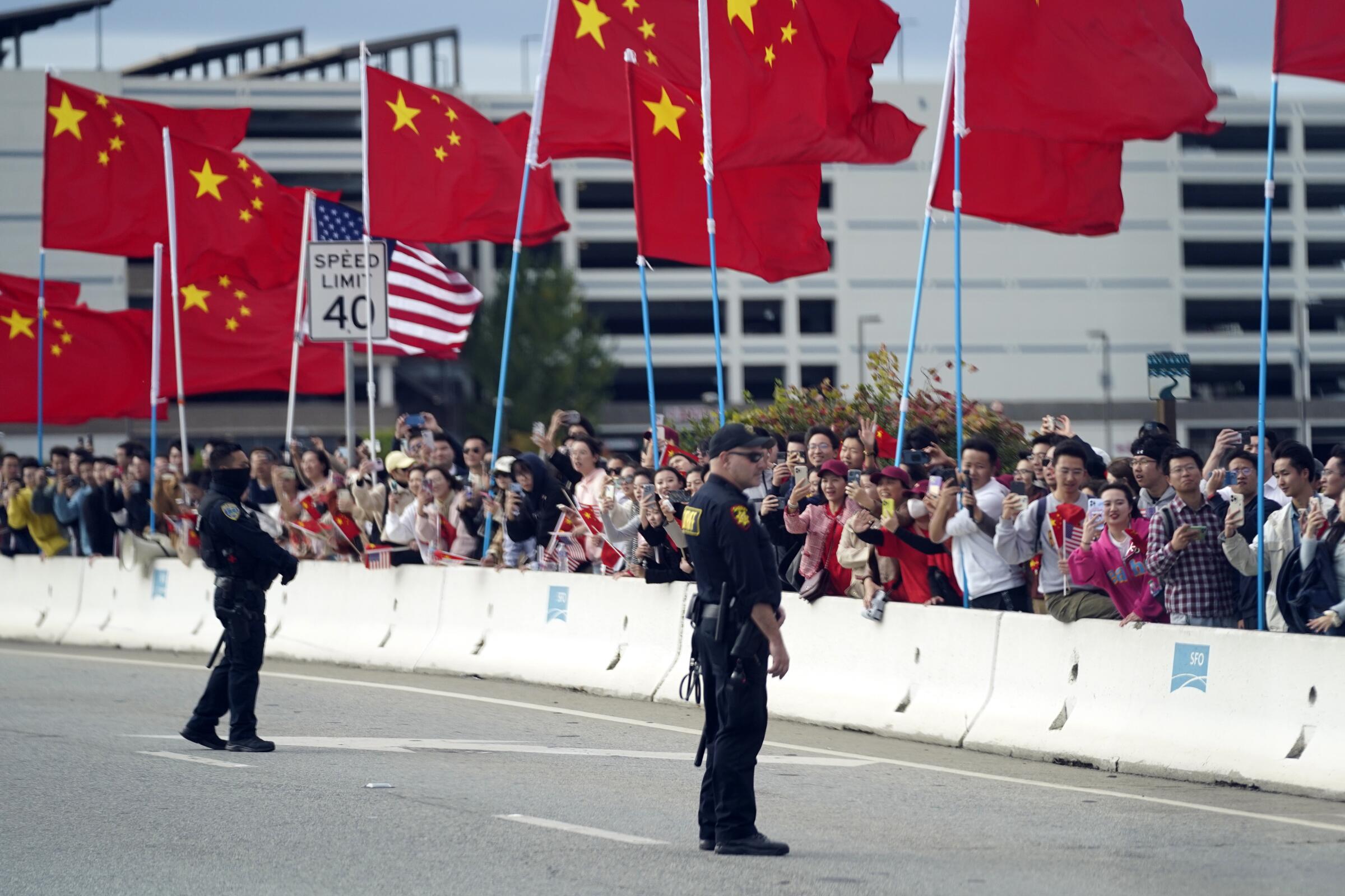 With American and Chinese flags flying, people watch on the side of a road.