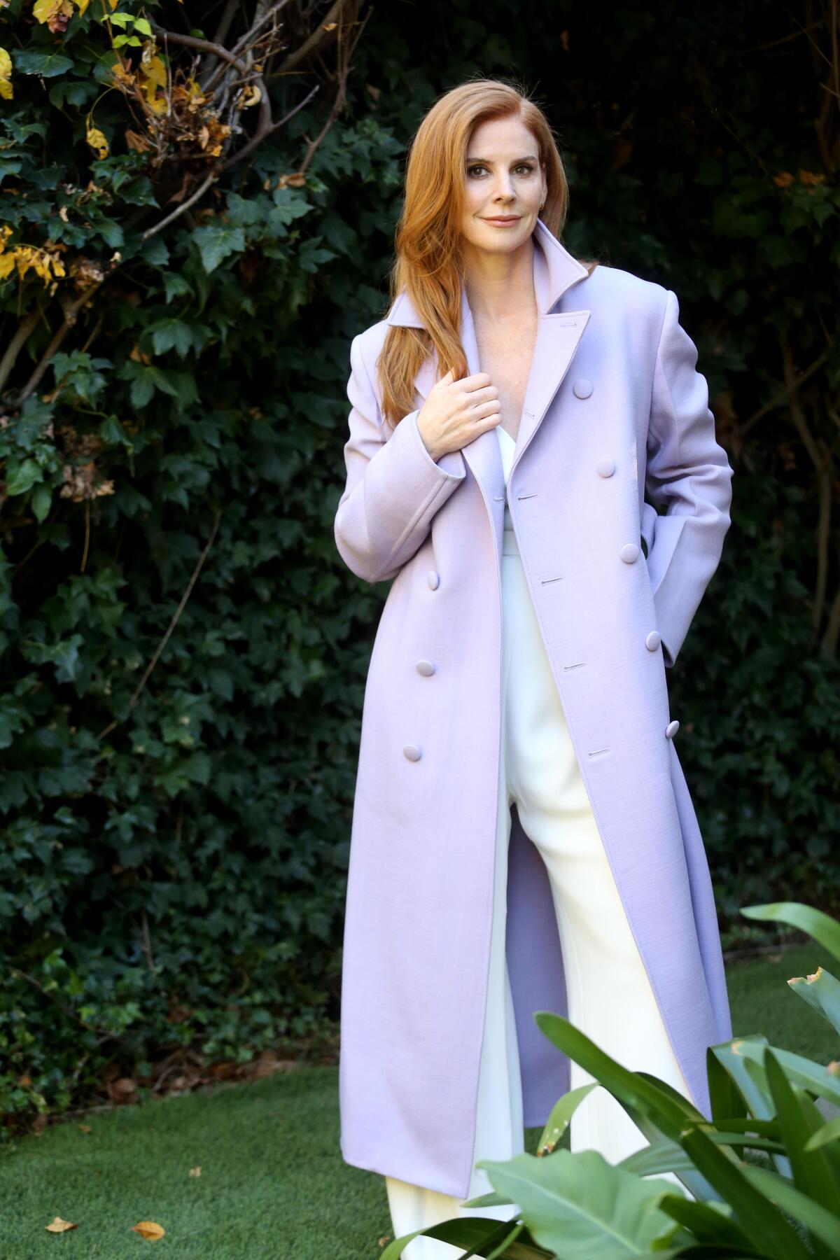 Actor Sarah Rafferty poses in a long lilac coat in her backyard.