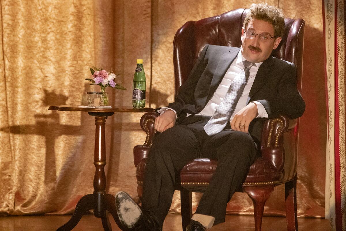 A man in a black suit sits in an armchair beside a table.