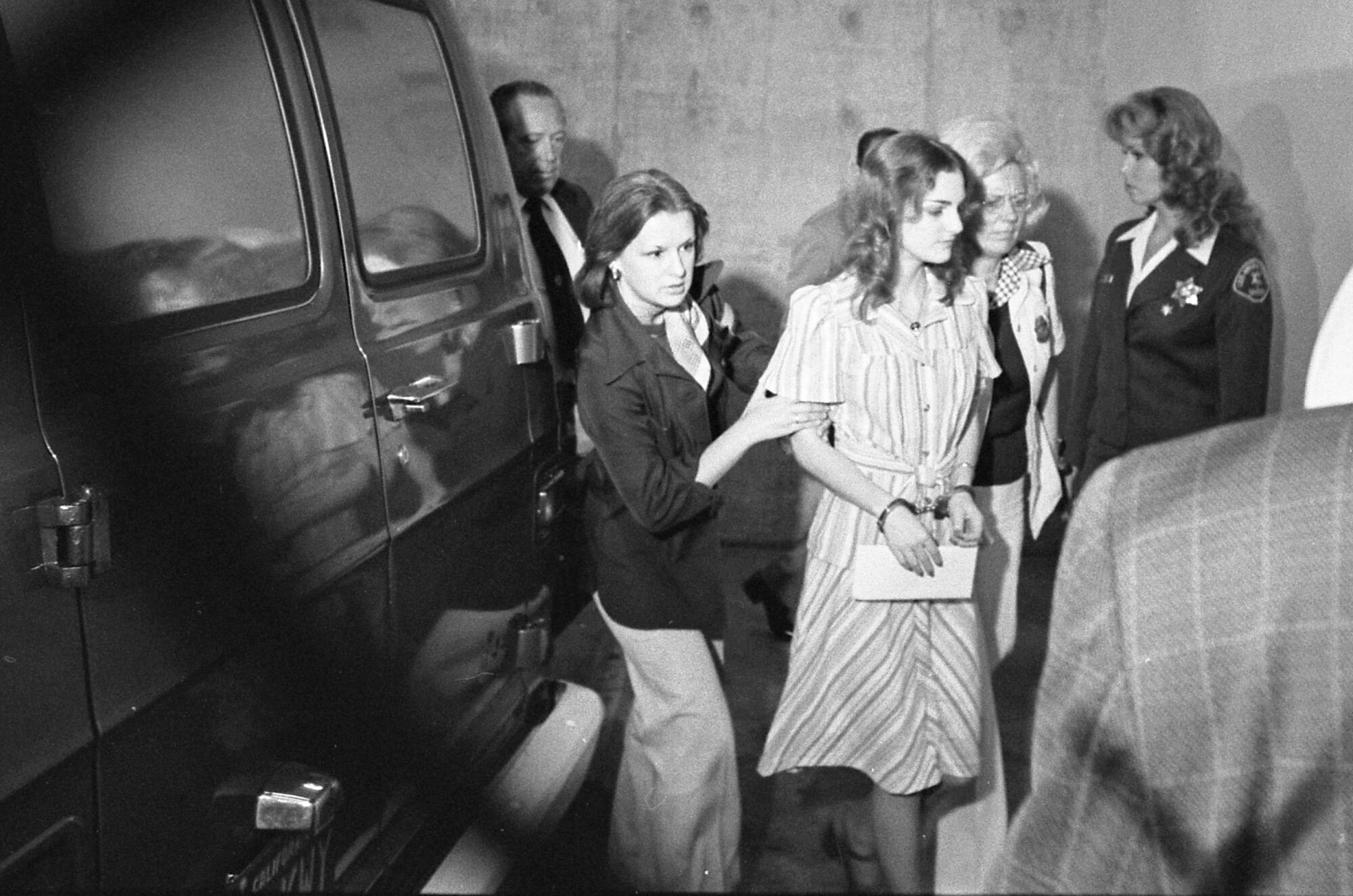 Patricia Hearst, wearing a striped dress and in handcuffs, is  escorted by two women at the inmate court entrance  