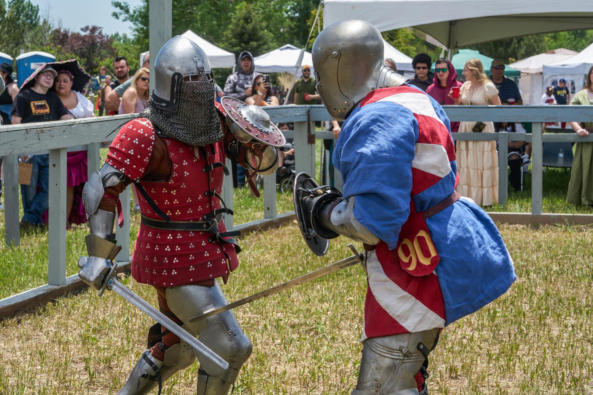 Two people in knight's armor, holding swords and shields, face off as a crowd watches 