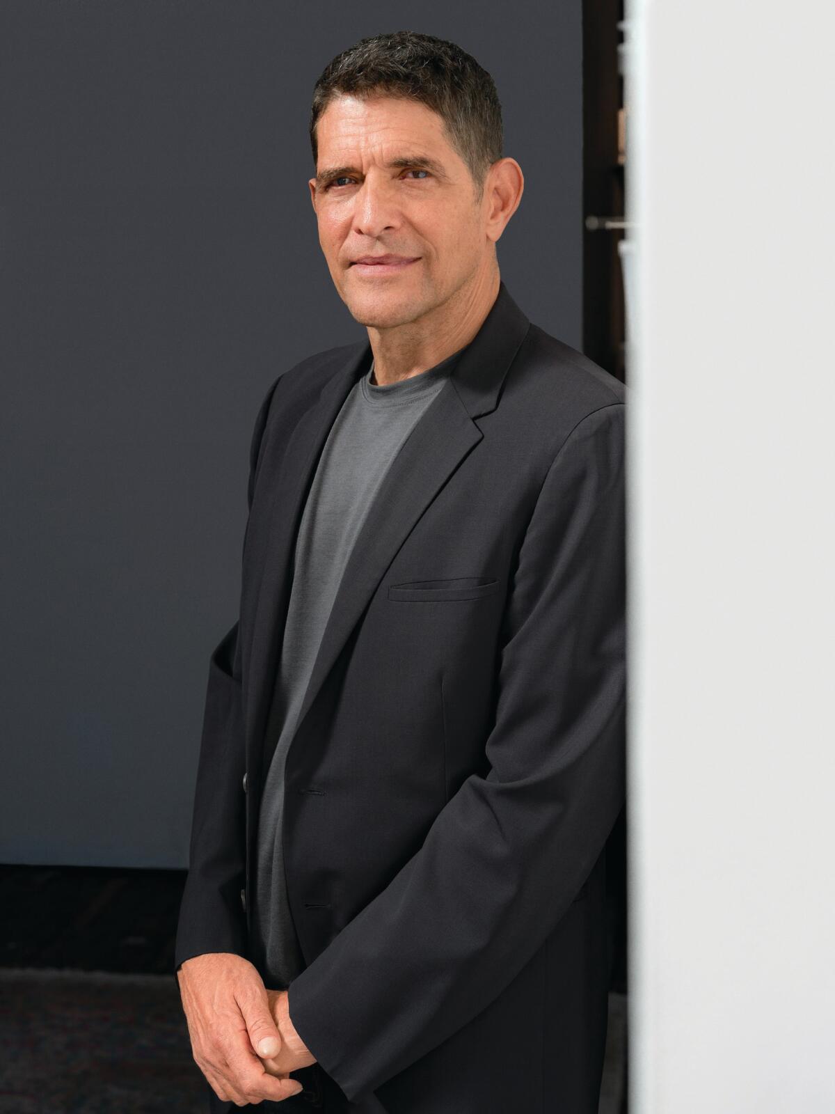 A dark-haired man in a dark blazer over gray T-shirt stands against a wall
