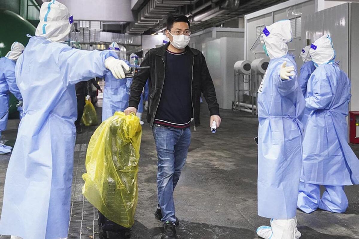 A man wheels a suitcase covered with a yellow plastic sheet past workers in protective suits, gloves and booties. 