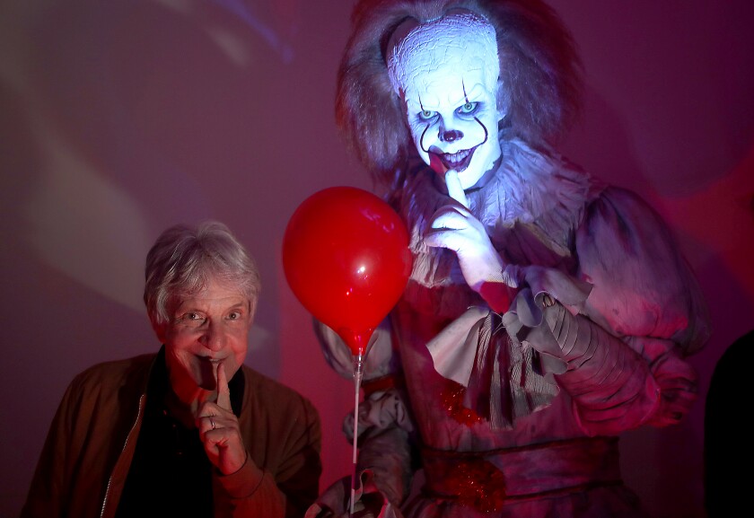 A man at left holds a finger to his lips. That's duplicated by a person to the right of a balloon with scary clown makeup.
