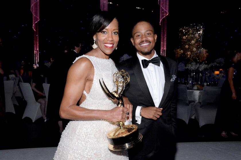 Regina King in a white gown holding an Emmy Award standing next to a young man with a mustache wearing a tuxedo