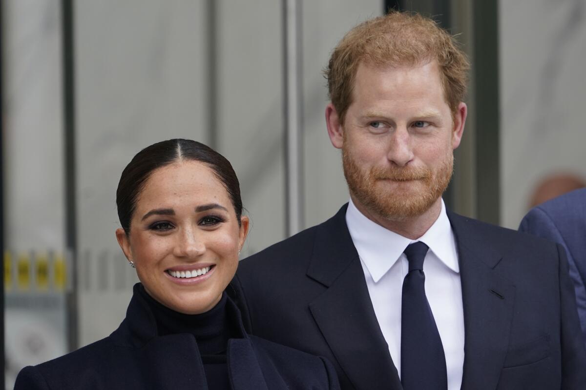 Meghan Markle in a black turtleneck with her hair in a bun standing next to Prince Harry in a suit