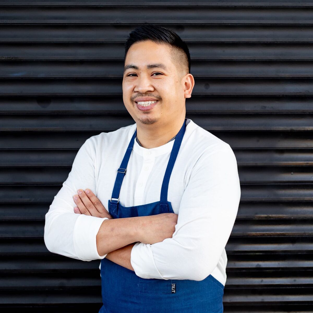 A man smiles, wearing an apron with his arms crossed