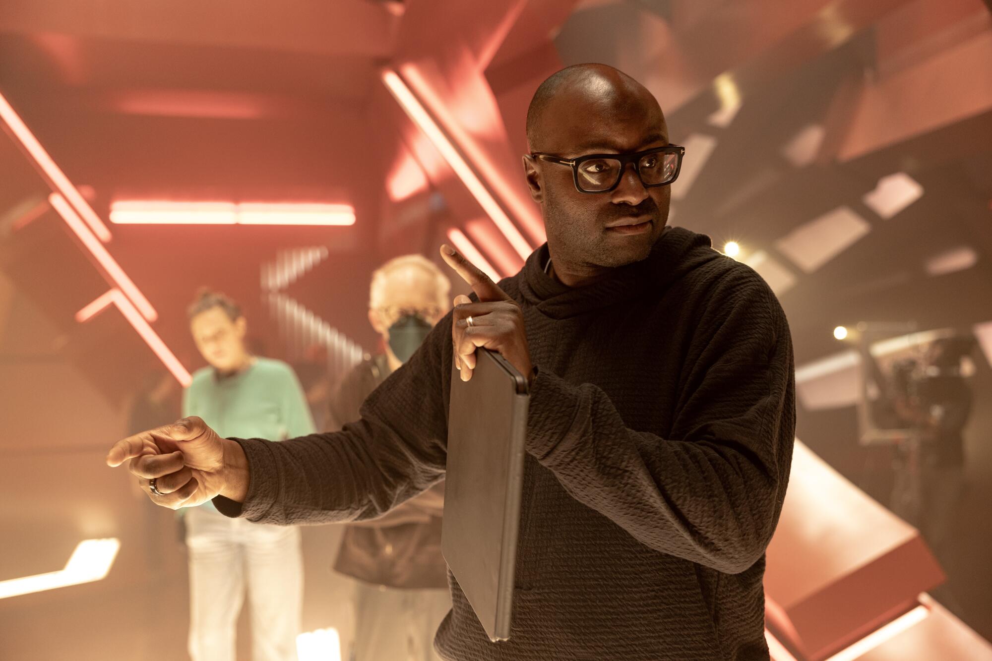 A bald man in a hoodie and black-rimmed glasses holding a computer tablet as he stands on a set.