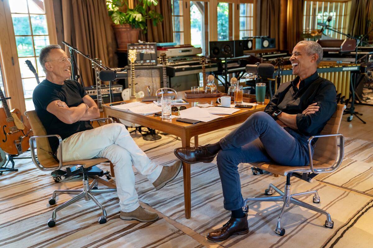 Springsteen and Obama sit at a table; in the background are guitars and recording equipment.
