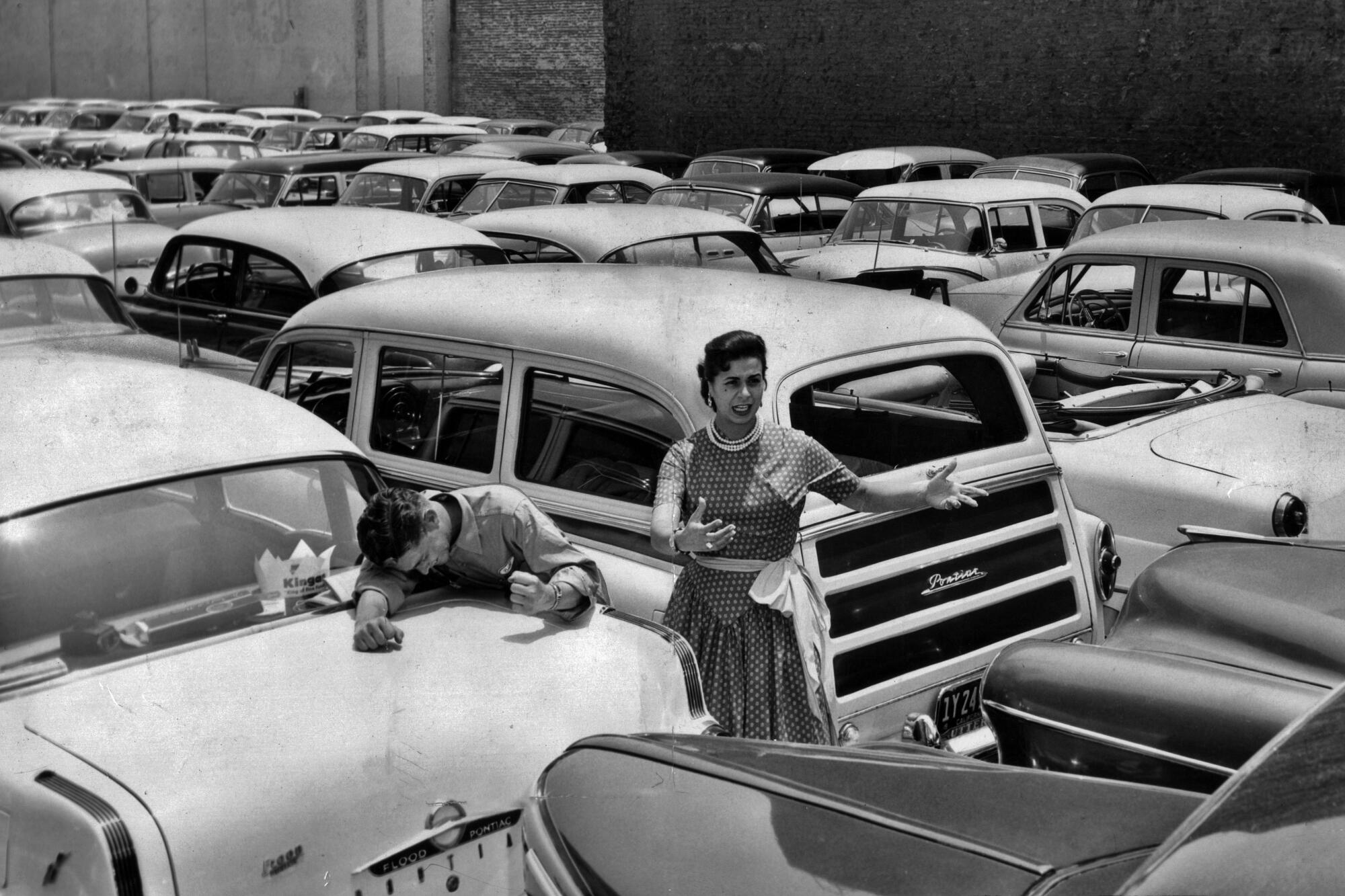 A black and white photo from the 1950s shows a woman standing in the middle of a large parking lot stuffed with cars.