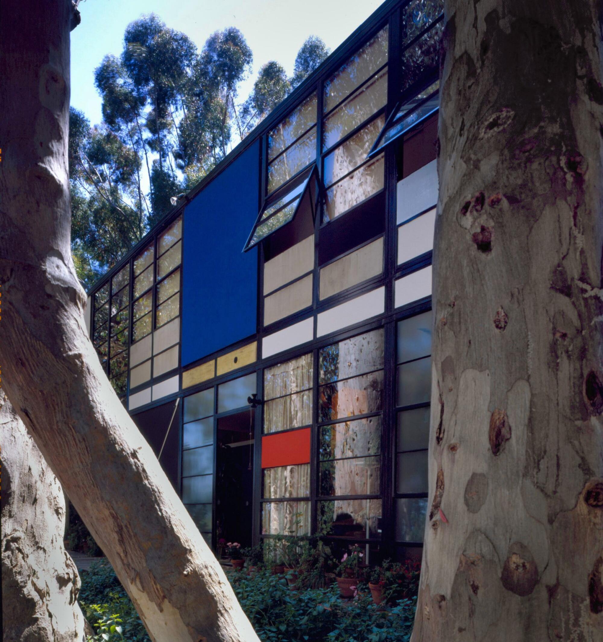 The Eames House facade — a grid of rectangular window panels, some splashed with color — is seen through trees.