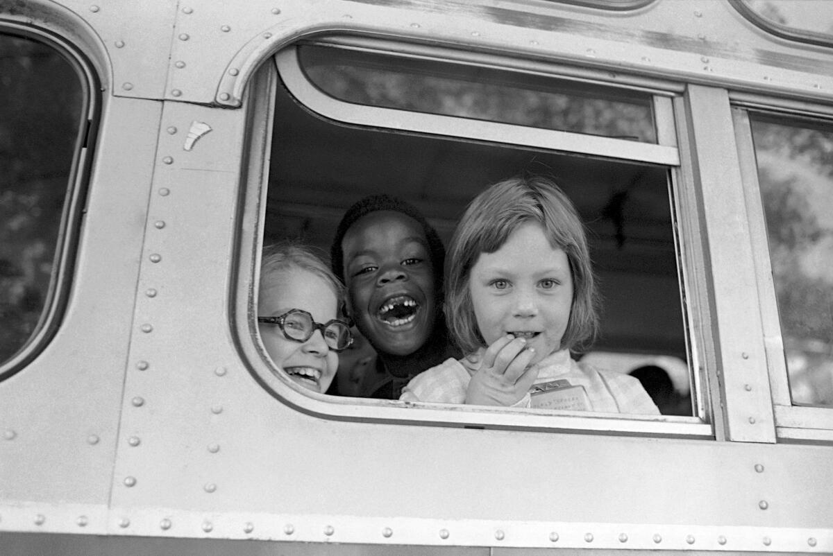 Three children -- one Black and two white -- smile from the window of a school bus
