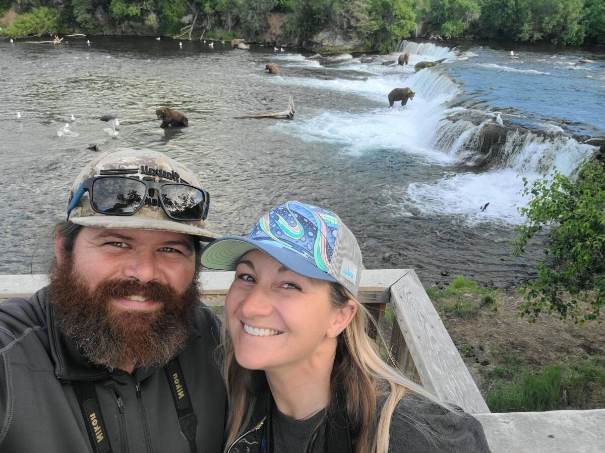 A bearded man and a woman with blond hair, both wearing caps, pose above a river, with bears in the background