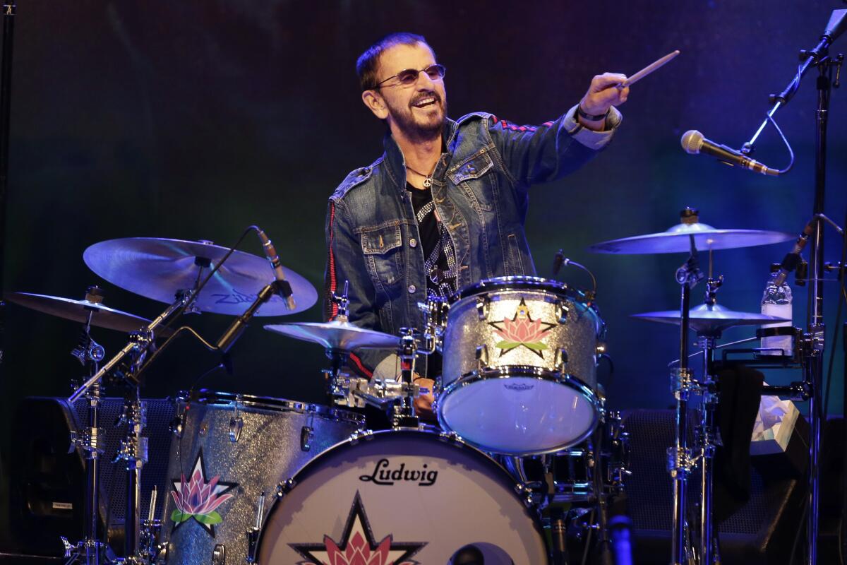 Ringo Starr sitting behind a drum kit  and pointing his left arm out